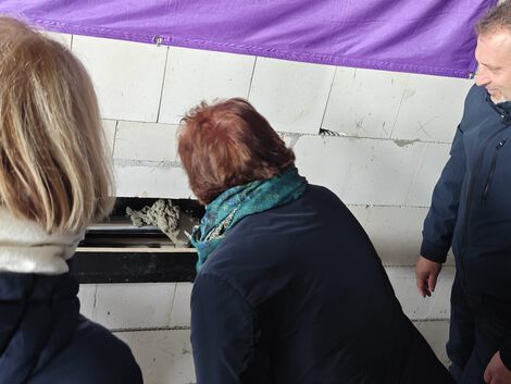 Drei Personen mit Jacken stehen vor einer Wand aus weißen Betonsteinen. Eine Person füllt ein Loch in der Wand mit Zement aus.