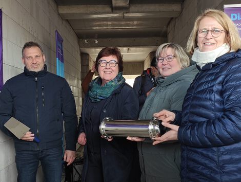 Gruppe von Personen in einem Gang, eine Person hält einen zylindrischen Metallbehälter